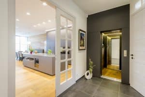 Flur einer Villa mit Homelift in schwarzer Verkleidung und Spiegel und Doppelflügeltüren aus weissem Holz mit Sprossen und Glaseinsätzen und einer Wohnküche mit Essbereich und Kochinsel von Planungsbüro Schaufler.