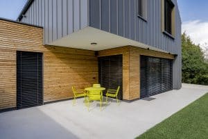 Einfamilienhaus mit überdachter Terrasse, gelbem Gartenmobiliar, mehreren Terrassentüren, Jalousien, Aussenspots und Fassadenelementen aus Holz von Planungsbüro Schaufler.
