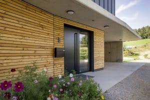 Hauseingang eines Einfamilienhauses mit einer schwarzen Eingangstüre mit Seitenlicht, Bau- und Fassadenelementen aus Beton, Blech und Holz mit Spots in der Betondecke von Planungsbüro Schaufler.