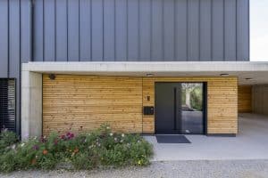 Hauseingang eines Einfamilienhauses mit einer schwarzen Eingangstüre mit Seitenlicht, Bau- und Fassadenelementen aus Beton, Blech und Holz mit Spots in der Betondecke von Planungsbüro Schaufler.