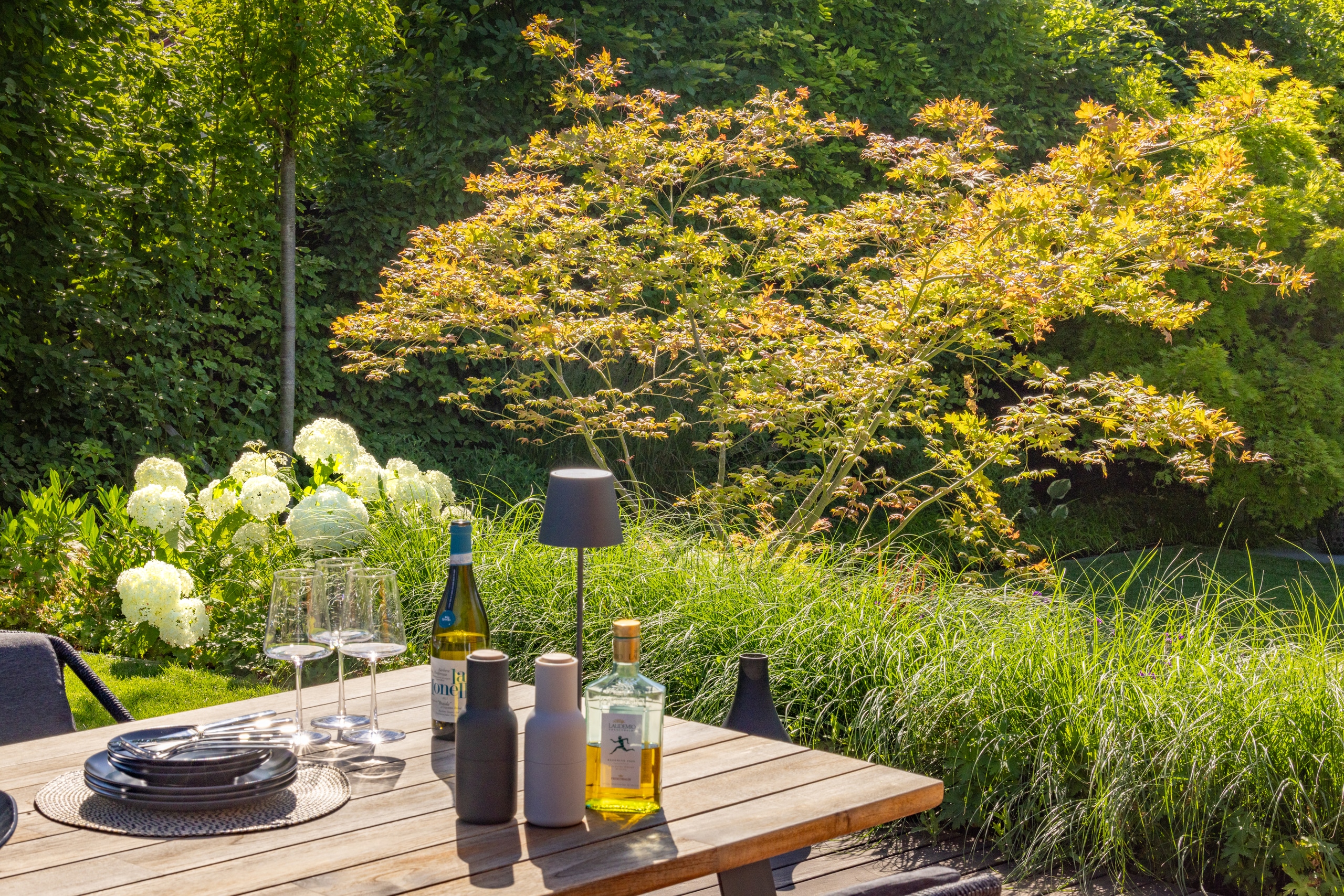 Praskac Pflanzenland Prachtgarten zeigt Garten mit Terrasse und Holztisch vor einem Beet mit Ziergräsern und Ahornbaum.