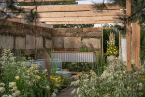 Praskac Pflanzenland Prachtgarten zeigt Blumenbeete und Sträucher vor einer Pergola mit Terrassenboden, Sitzbank und Steinmauer.