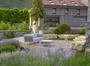 Prachtgarten zeigt einen Natursteingarten, mit Outdoorsofa und Pouf, Steinschlichtung als Bezäunung, Steinstufen, Steinblöcke zum Sitzen, Bäumen, Heckenkugeln und blühenden Sträuchern, Wildblumen und Lavendel.
