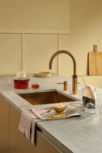 Eine moderne Kücheninsel mit Marmorarbeitsfläche, hellbraunen Küchenfronten, passenden Wänden, einer quadratischen Spüle und einer Quooker Fusion Armatur mit C-Auslauf in "Messing Patina" mit passendem Einbau-Seifenspender.