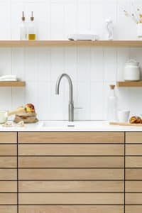 Eine moderne Küche mit hellen Holzfronten, passenden Schweberegalen, weisser Arbeitsfläche und weissen Stabfliesen mit einer Quooker Fusion-Armatur mit C-Auslauf in Chrom aus rostfreiem Edelstahl.