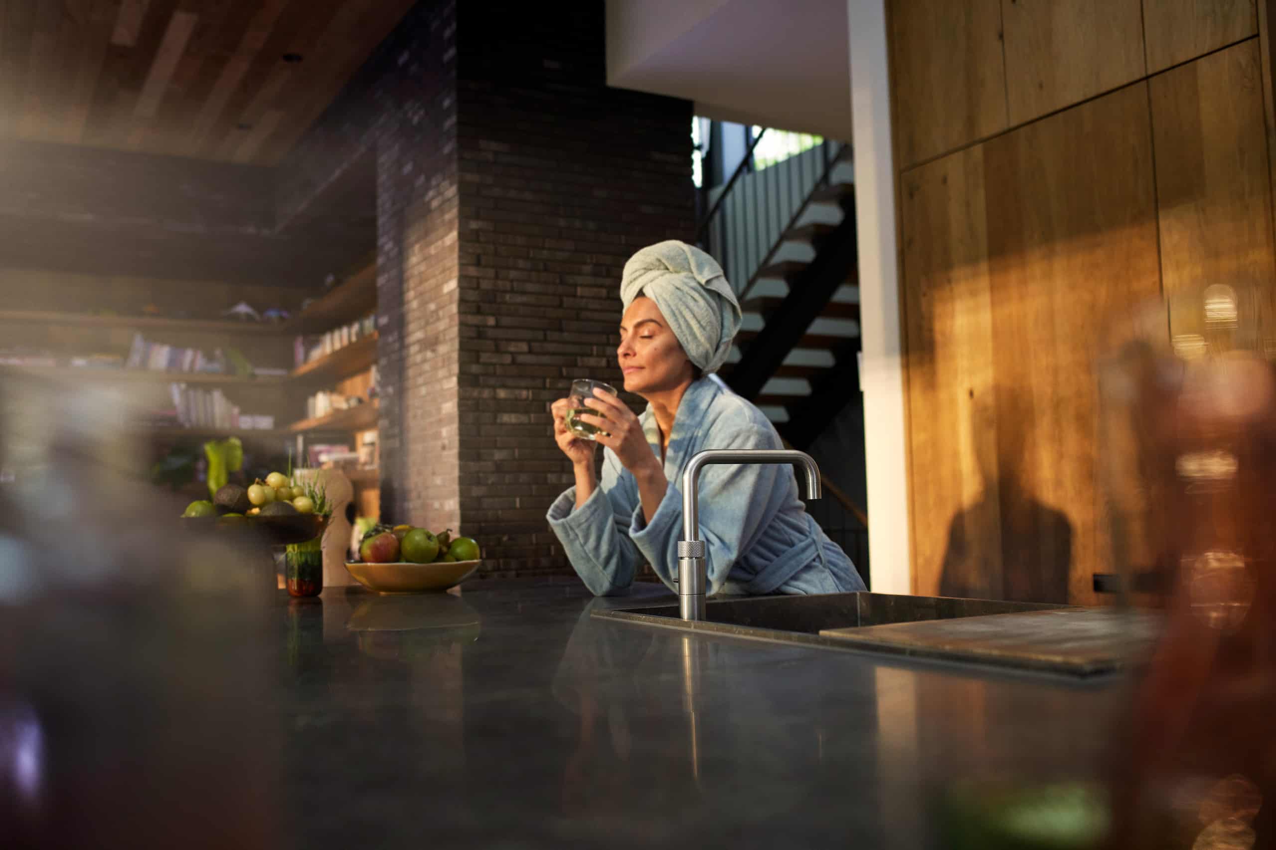 Eine Frau in Bademantel und Handtuchturban genießt ein Glas Wasser aus modernen Quooker-Armatur im Square-Design in einer Villa mit stilvoller offener Küche von Quooker.