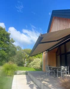 Ein Haus mit Satteldach und Holzfassade, gemauertem Terrassenpodest und einer Gelenkarmmarkise mit schwarzem Rahmen und gold-beigem Markisenstoff in Seitenansicht von Sonnenschutz Rachbauer.