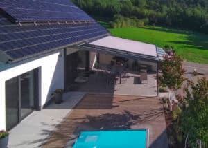Ein Haus mit Solardach, Garten, Pool und einem überdachten gefliestem Terrassenbereich mit Pergola bzw. Sommergarten mit geneigtem Glasdach und beiger Glasdachmarkise von Sonnenschutz Rachbauer.