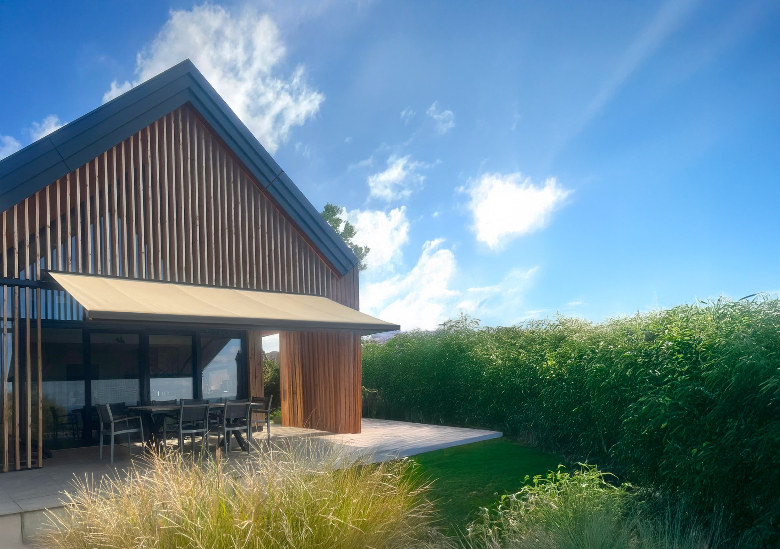 Ein Haus mit Satteldach und Holzfassade, gemauertem Terrassenpodest und einer mit der Hausfassade farblich abgestimmten Gelenkarmmarkise mit schwarzem Rahmen und gold-beigem Markisenstoff von Sonnenschutz Rachbauer. Header MONTAGE