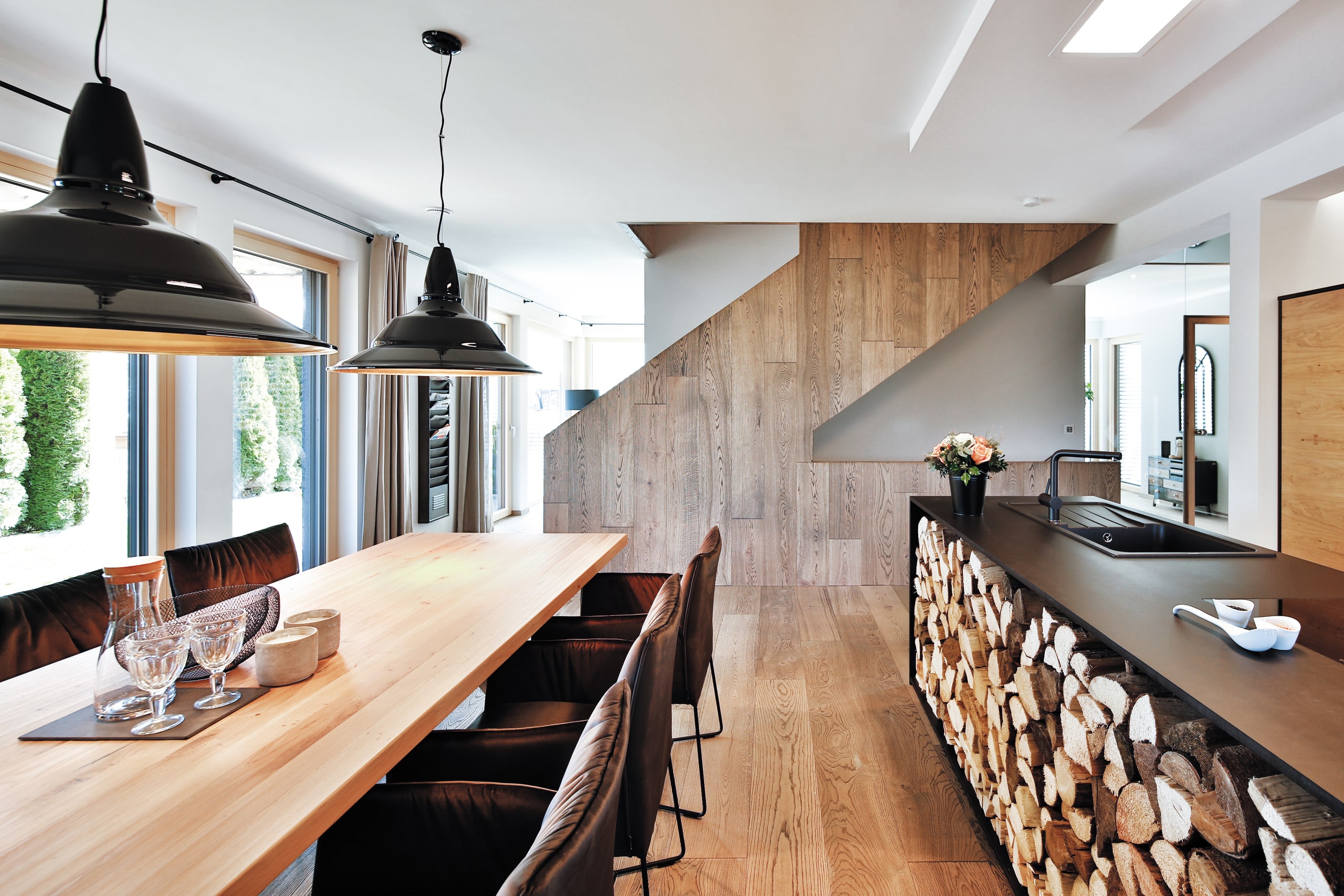 Einfamilienhaus Heidi mit Kücheninsel mit integriertem Holzlager, Essbereich und Blick auf die holzverblendete Seitenwand des Treppenaufgangs von Regnauer Fertighaus.