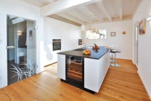 Küche mit Kücheninsel, Tresen, Barhockern, einem Weintemperierer, Glastür zum Vorzimmer und heruntergesetzter Holzdecke mit Balken von Regnauer Fertighaus.