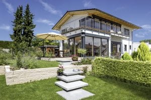Einfamilienhaus mit Satteldach, halbüberdachter Eckterrasse mit mehreren Terrassenzugängen, einem Eckbalkon mit Eisengeländer im Obergeschoß und einem Garten mit schwebenden Steinfliesen von Regnauer Fertighaus.