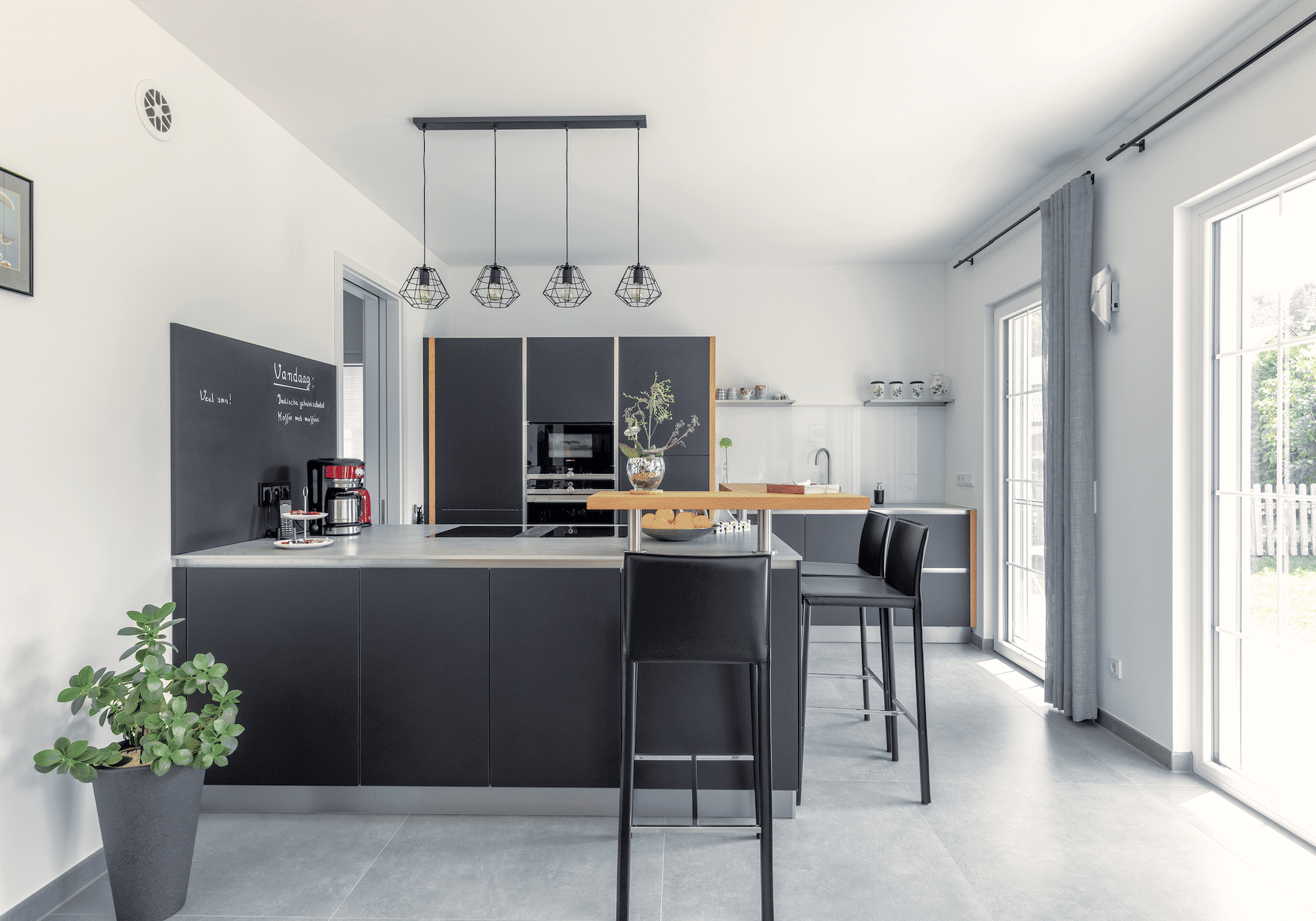 Regnauer zeigt eine schwarze Küche mit einer grauen Arbeitsplatte, Hochstühlen und einer Bar aus Holz und Glastüren mit grauem Vorhang und Zugang in den Garten.