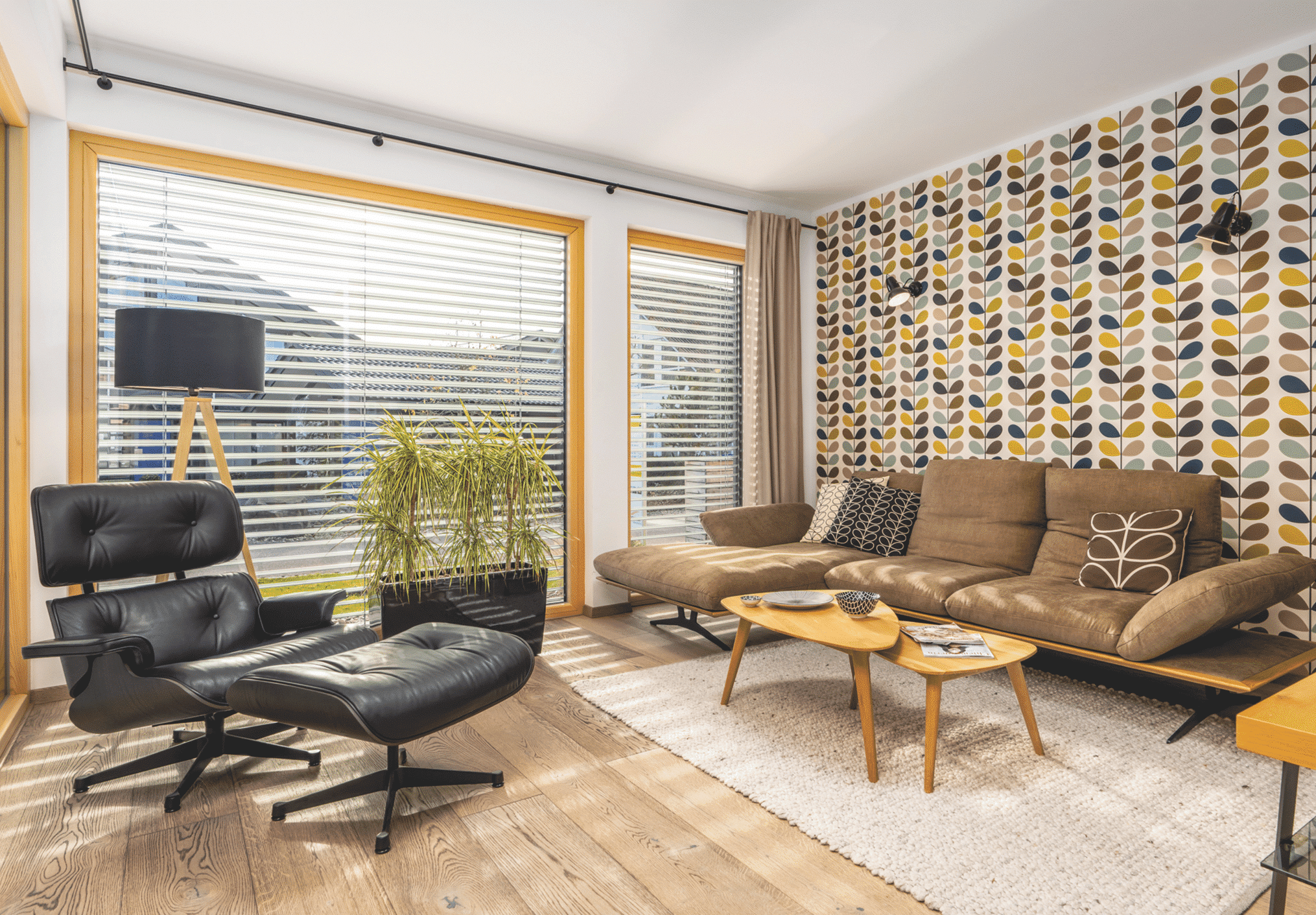 Regnauer zeigt ein Wohnzimmer mit gemusterter Tapete, einer braunen Ledercouch und einen schwarzen Ledersessel mit Hocker und Zugang zum Balkon.