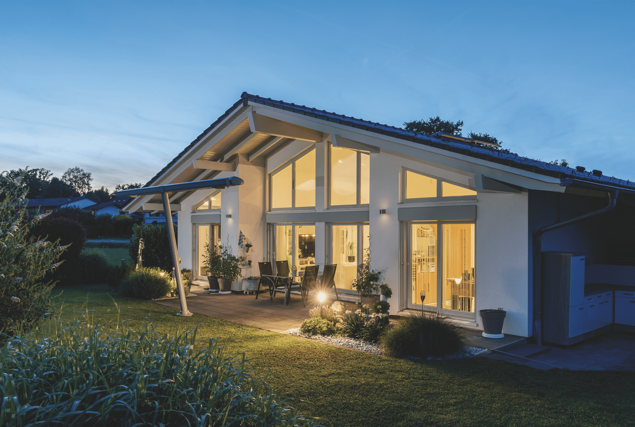 Regnauer zeigt ein weisses Einfamilienhaus  im Bungalow Stil mit großen Glasfronten und einer gefliesten Terrasse mit einer Sitzgruppe und warmer Beleuchtung.