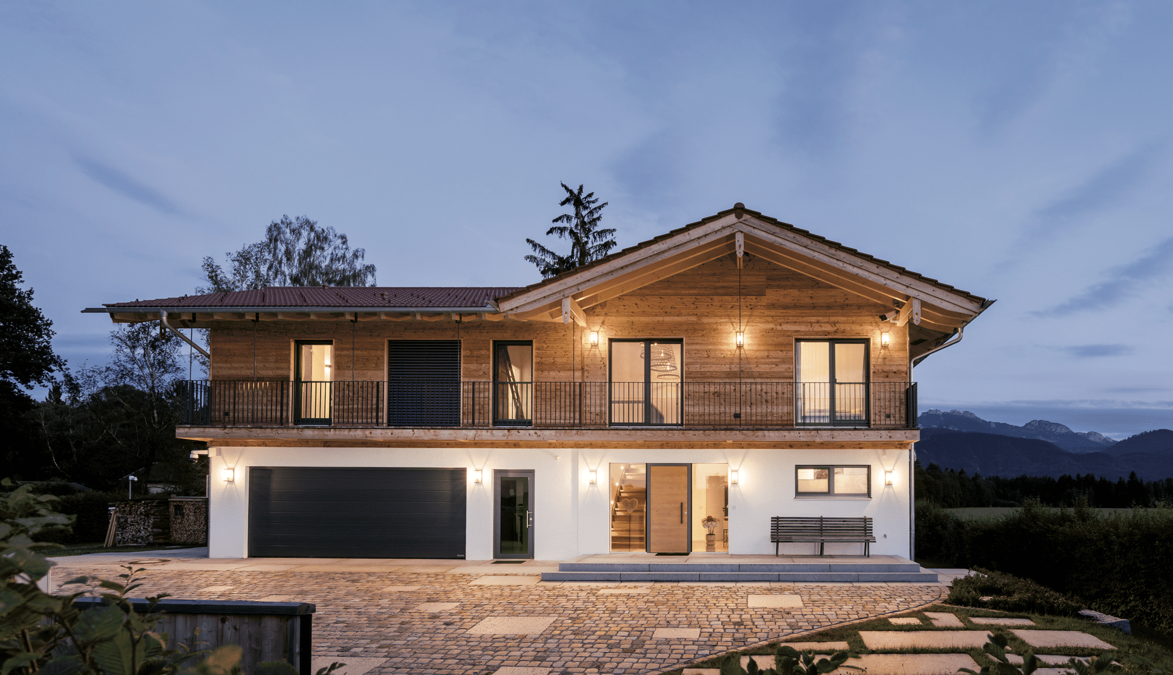 Regnauer zeigt ein Einfamilienhaus mit heller Holzverkleidung und einer Doppelgarage mit dunklem Garagentor und einen Balkon mit Metallgeländer.