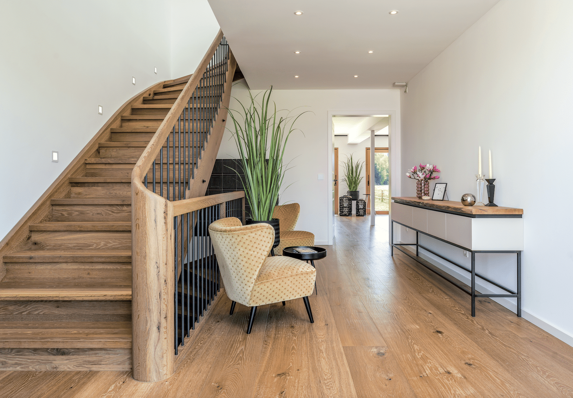 Regnauer zeigt einen Vorraum mit Holzboden und gemütlichen hellen Ohrensesseln, einem runden, schwarzen Beistelltisch und Holztreppen in das nächste Stockwerk.