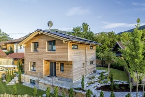 Regnauer Fertighaus zeigt Holzhaus mit Satteldach, Fenster mit dunkelgrauem Rahmen, nach innen versetztem Hauseingang auf einem Ziegelsteinsockel mit großem Steingarten.