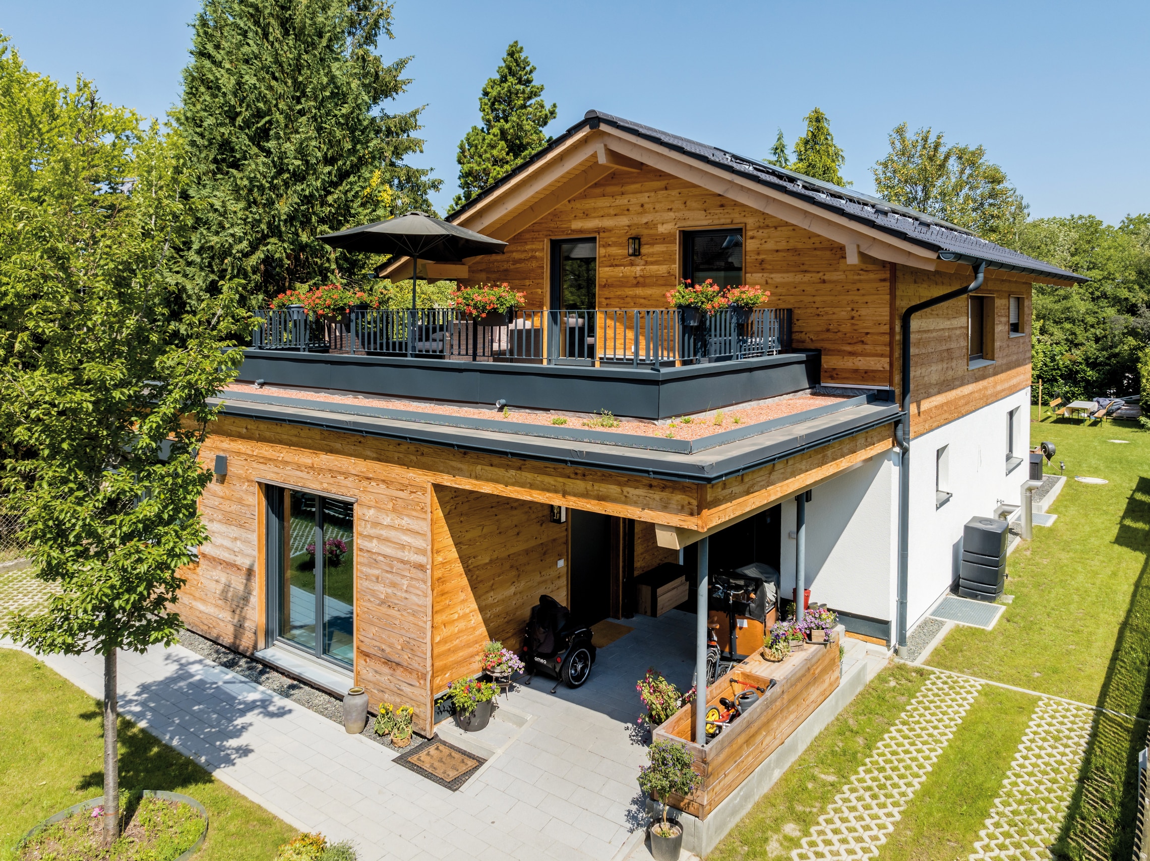 Regnauer Fertighaus zeigt modernen Holzhaus mit Satteldach mit nach innen versetztem Eingangsbereich und einer großen Terrasse mit dunkelgrauem Geländer und Schotterumrandung, die begrünt werden kann..