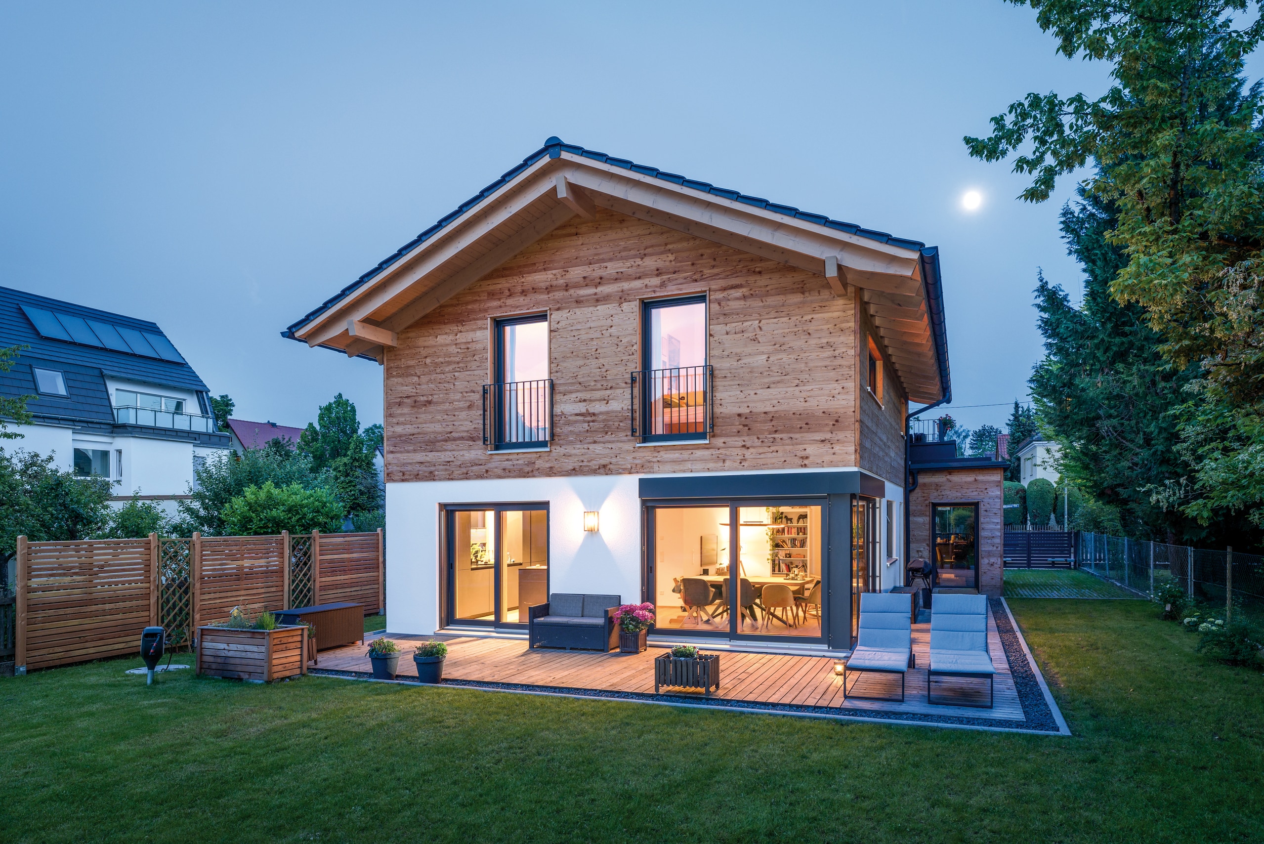 Regnauer Fertighaus zeigt elegantes Einfamilienhaus mit Satteldach, holzverkleideter Fassade, großen Glasfronten im Erdgeschoss und Terrasse bei Abendstimmung.