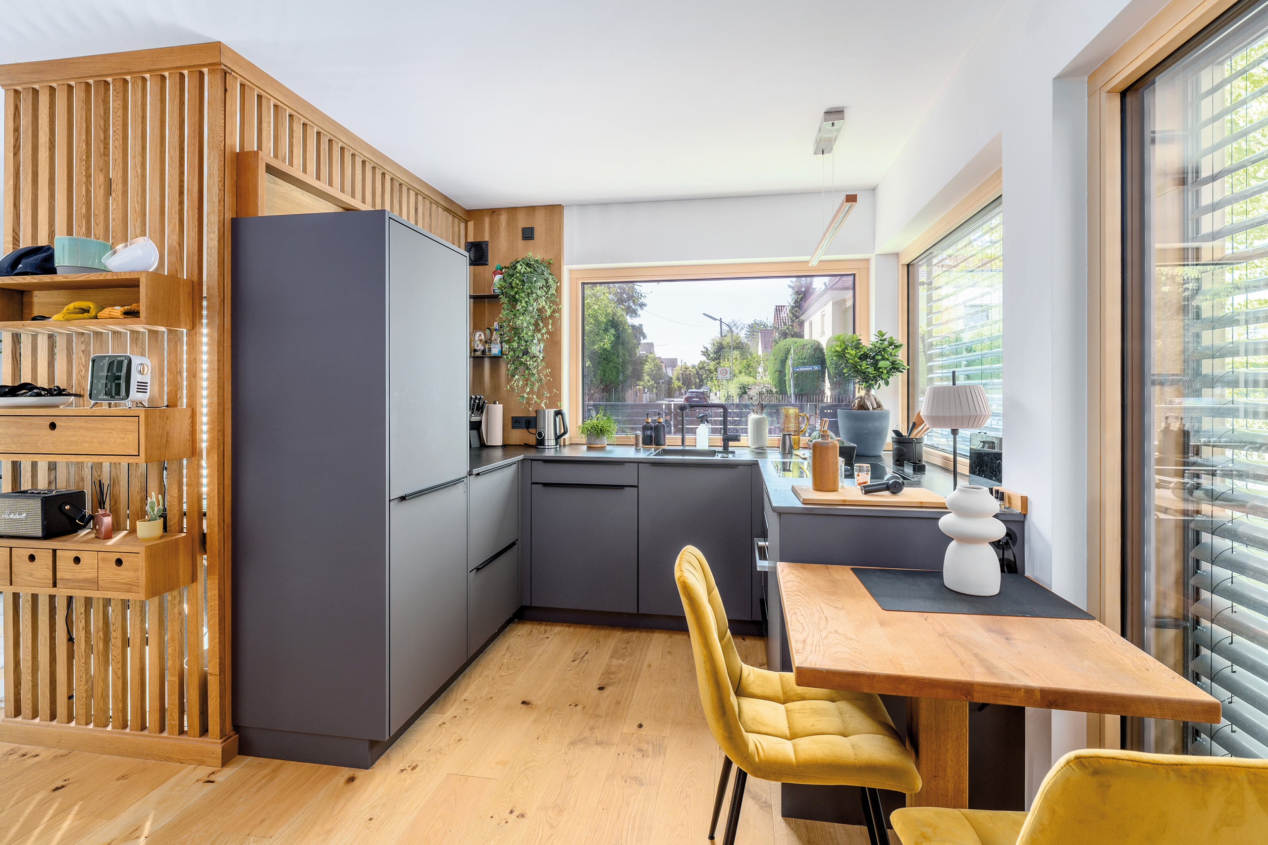 Regnauer Fertighaus zeigt Küche mit großem fixverglastem Fenster, dunkelgrauen Fronten und einem angebauten Esstisch mit zwei Stühlen und Raumtrenner mit Holzsprossen verkleidet mit Ablageflächen.