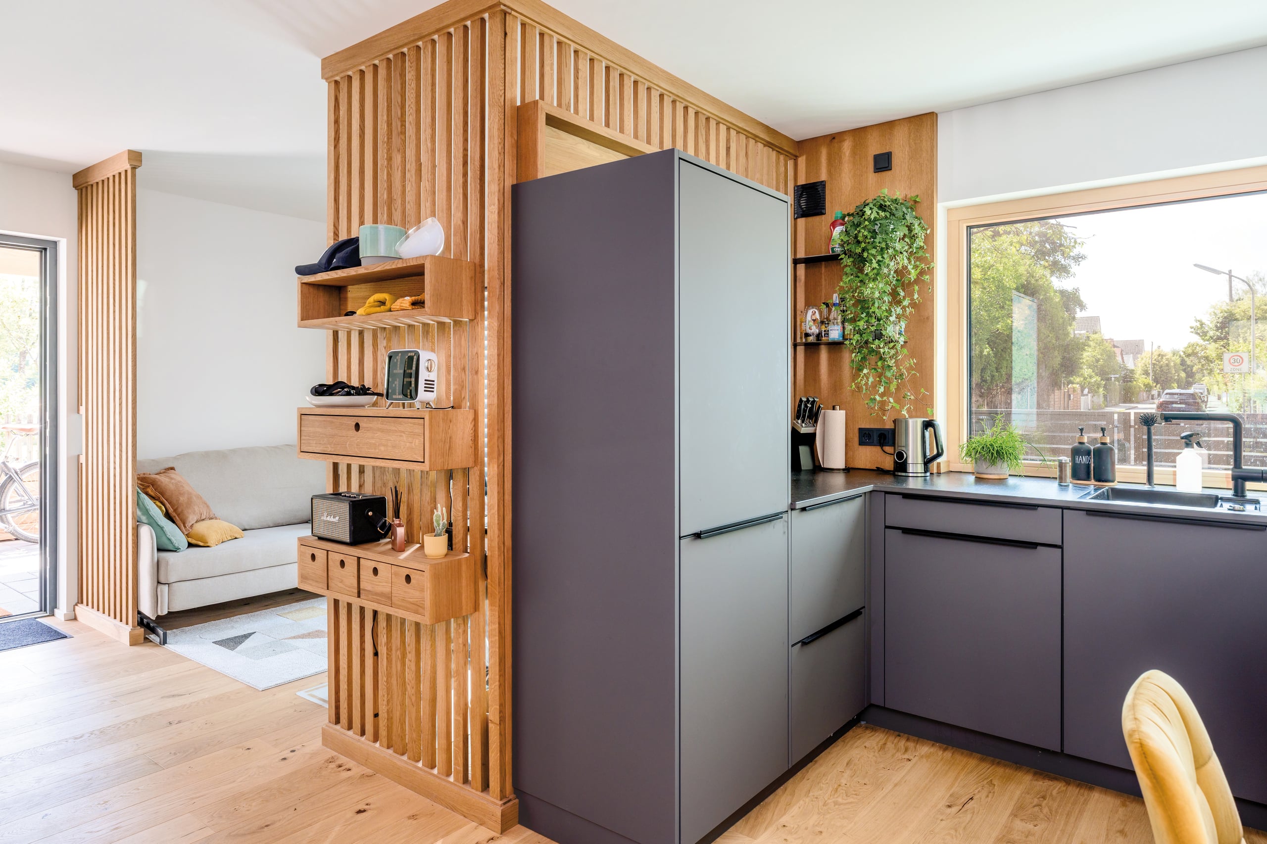 Regnauer Fertighaus zeigt Küche mit großem fixverglastem Fenster, dunkelgrauen Fronten an einem Raumtrenner mit Holzsprossen verkleidet und Ablageflächen mit Blick in das Wohnzimmer.