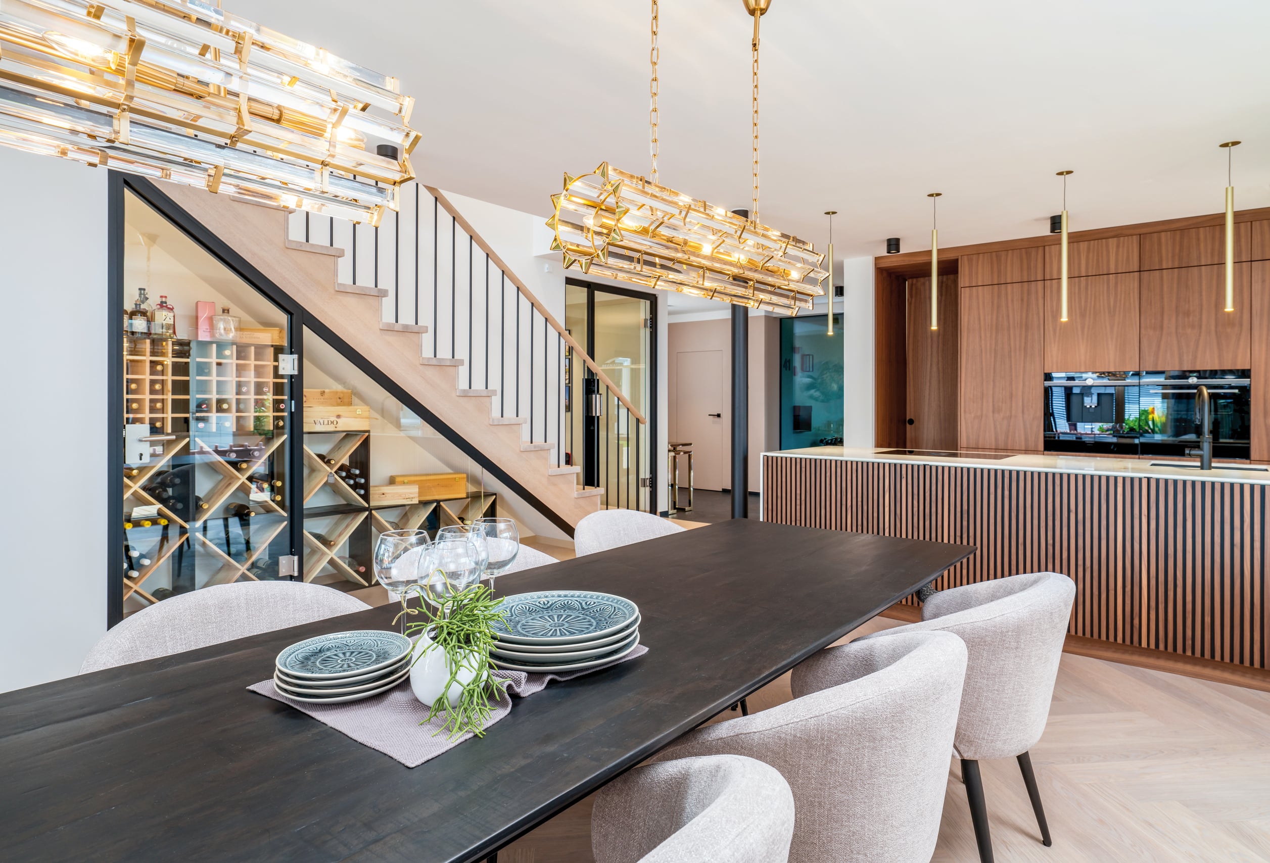 Regnauer Fertighaus zeigt Essbereich mit extravaganten Leuchten, großem Holztisch, stoffbezogenen Stühlen mit Blick auf die Küche mit Holzfronten und Stiegenaufgang mit Treppenunterbau, der als Weinregal dient.