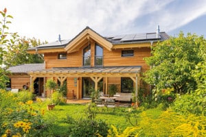 Regnauer Fertighaus zeigt Einfamilienhaus mit Zwerchdach, überdachter Terrasse aus Holz mit Holzsäulen mit Blick in den Garten.