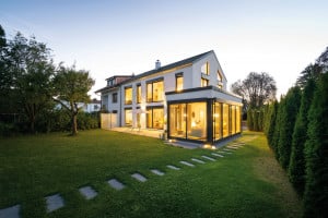 Regnauer Fertighaus zeigt Einfamilienhaus mit Satteldach, angebauten Wintergarten mit großen Fensterflächen, Terrasse und Steinplatten-Weg in den Garten bei Abendstimmung.
