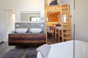 Ein großzügiges Dachgeschoss‑Badezimmer mit Dachschräge, schwebendem Doppelwaschtisch aus dunklem Holz, aufgesetzten Waschschüsseln, wandmontierten Armaturen, einer freistehenden Badewanne, einem gläsernen Sauna‑Abteil und integrierter Leiter zu einem Stauraumbereich von Regnauer.