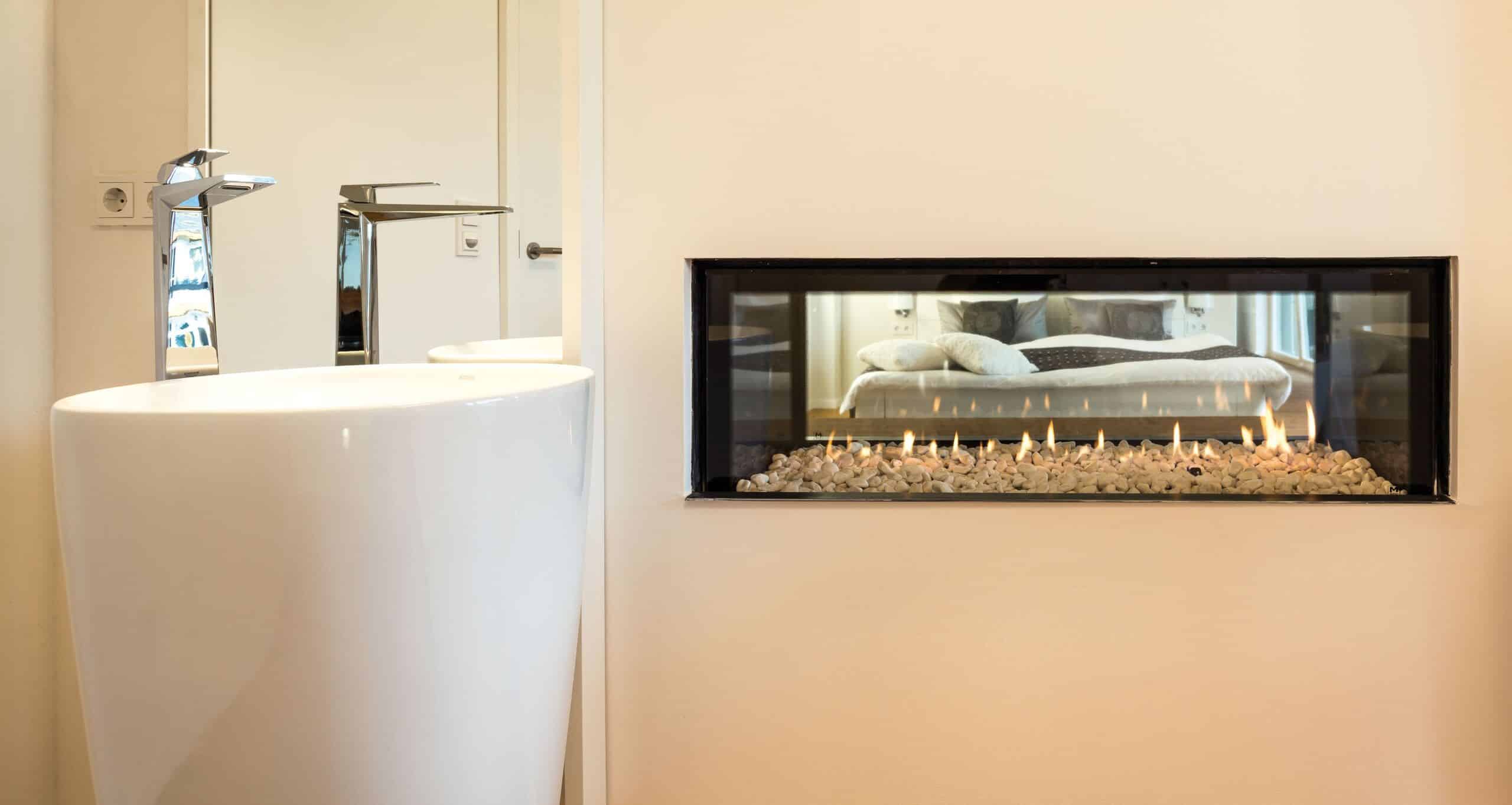 Ein edles Badezimmer mit bodentiefen Waschsäulen und einem Tunnelkamin mit Steinen, der durch die Wand einen Blick ins angrenzende Schlafzimmer ermöglicht, von Regnauer.