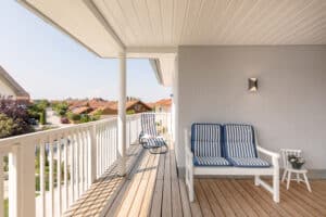 Ein klassisch elegantes Vitalhaus mit überdachter Veranda mit weisser Holzdecke, Holz‑Terrassendielen, Balkon und Holzgeländer, das Wohnraum und Außenbereich harmonisch verbindet von Regnauer Hausbau.