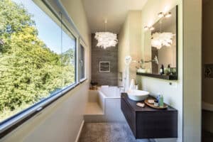 Ein modernes längliches Badezimmer mit erhöhter gemauerter Badewanne, Fensterfront, Waschtisch und Naturblick von Regnauer.