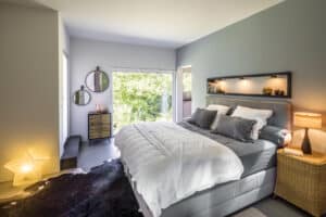 Ein modernes Schlafzimmer mit bodentiefem Glaselement, Terrassentür, Durchgang zum angrenzenden Raum, Polsterbett und hellem, offenem Wohnkonzept von Regnauer.