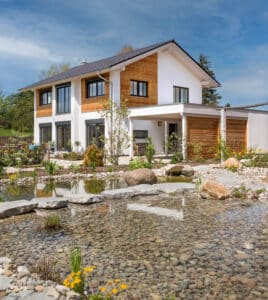Einfamilienhaus "Glonn" mit Holzfassadenelementen, überdachtem Eingangsbereich, Terrassentüren mit schwarzem Alurahmen, gepflastertem Außenbereich und Schwimmteich von Regnauer.