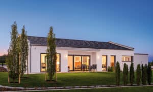 Ein moderner Bungalow mit Satteldach, mehreren Terrassenschiebetüren, überdachter Terrasse und Gartenfläche bei Sonnenuntergang von Regnauer.