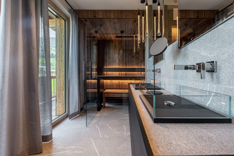 RUHA präsentiert eine Badezimmer mit kompakter Sauna aus dunklem Holz und einem Doppel-Waschtisch mit aufgesetzten Waschbecken aus Glas.