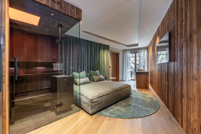 RUHA präsentiert einen Homespa-Raum mit kompakter Sauna aus dunklem Holz, einer bettähnlicher Couch und einem Holzschrank mit aufgesetztem Waschbecken.