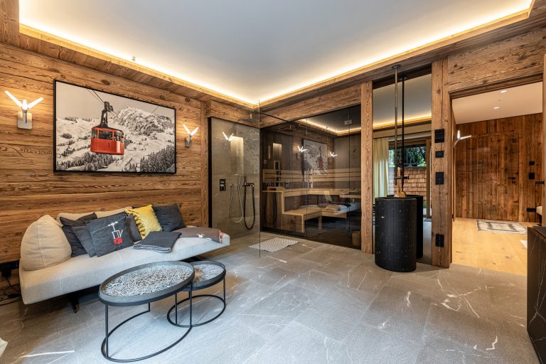 RUHA präsentiert einen Homespa-Raum mit großer Sauna mit Rauchglas und aus Altholz, einer gemütlichen Couch und einem stehenden Waschbecken.