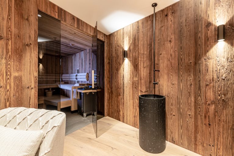 RUHA präsentiert einen Homespa-Raum mit großer Sauna mit Rauchglas und aus Altholz und einem stehenden Waschbecken.