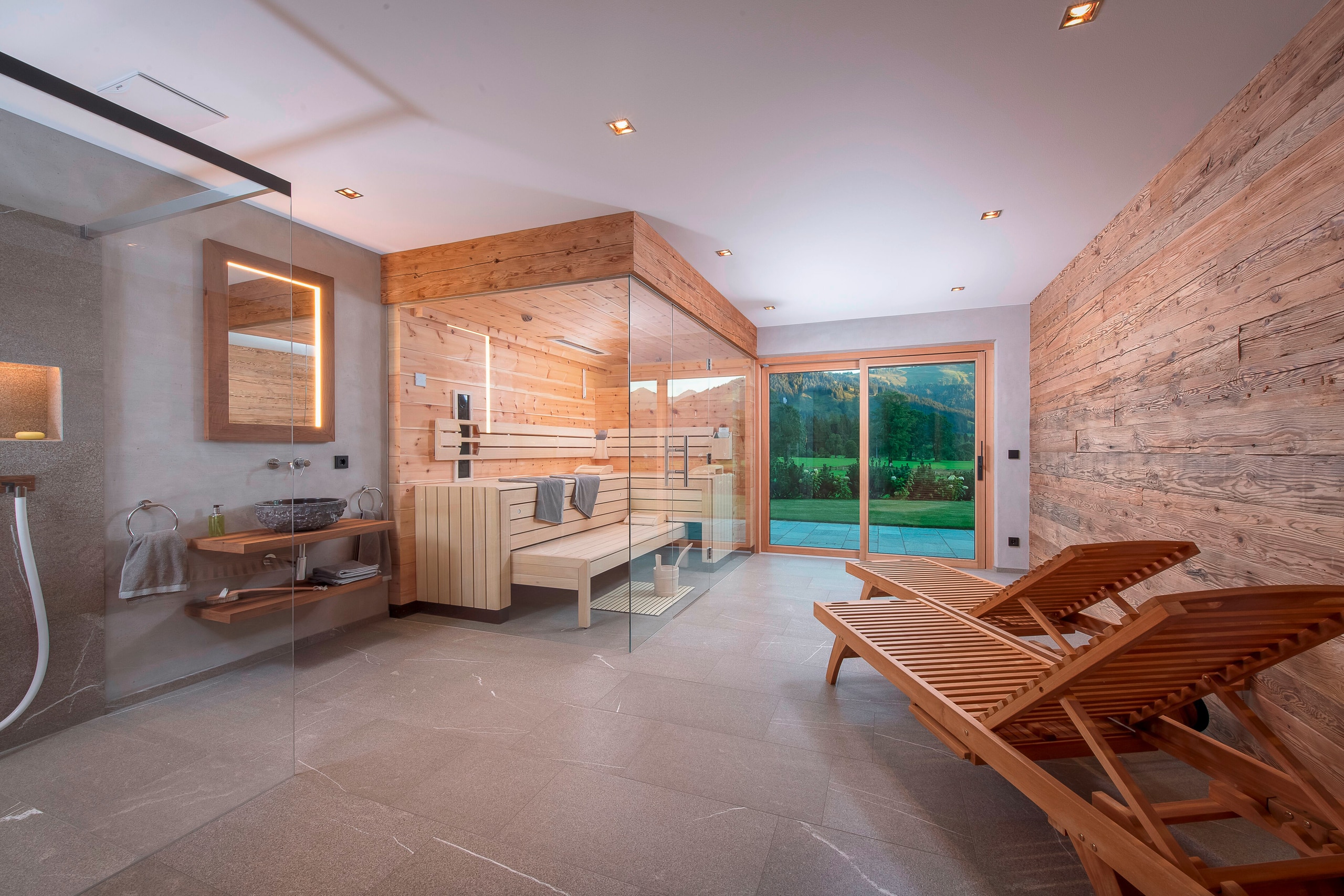RUHA präsentiert einen Homespa-Raum mit großer Sauna aus Altholz, einem Waschbereich, einer barrierefreien Dusche und zwei Holz-Liegestühle mit Zugang auf die Terrasse.