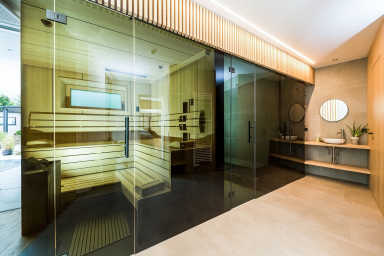 RUHA präsentiert eine große Sauna aus hellem Holz und Rauchglas vor einem Waschtisch mit freistehendem Waschbecken.