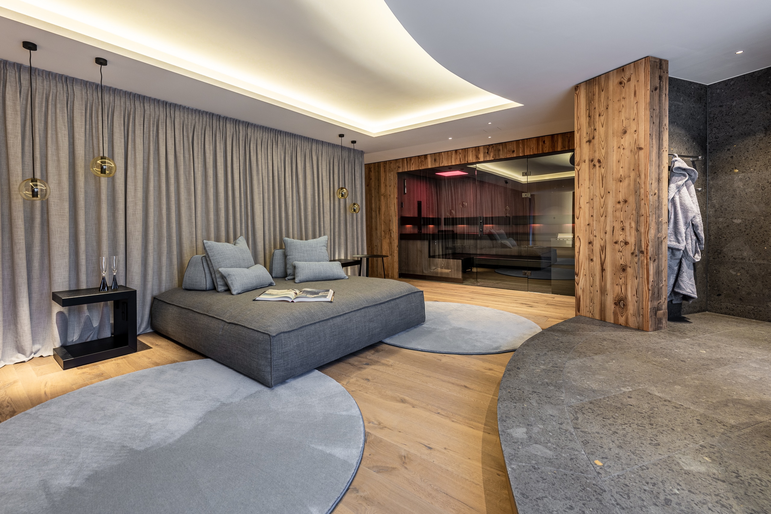 RUHA präsentiert einen Homespa-Raum mit bettähnlicher Couch, einer gefliesten Duschnische und einer großen Sauna mit Altholzverkleidung und Rauchglas.