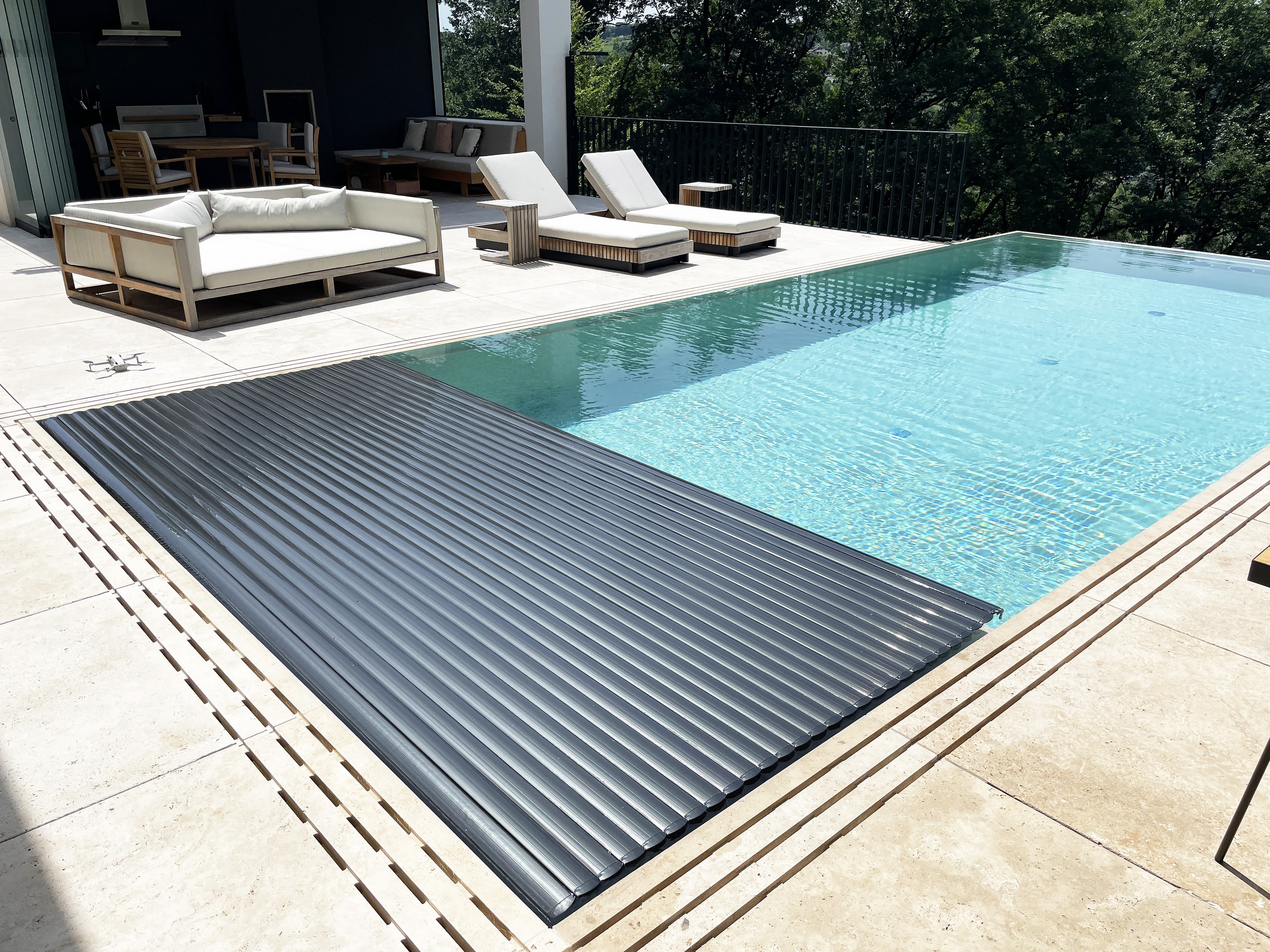 Ein Infinity‑Pool mit Überlaufrinne und Fliesen‑Poolumrandung, Relaxliege, halboffener schwarzer Poolabdeckung und schwarzem Sprossen‑Terrassengeländer von Ruha Stelzmüller.