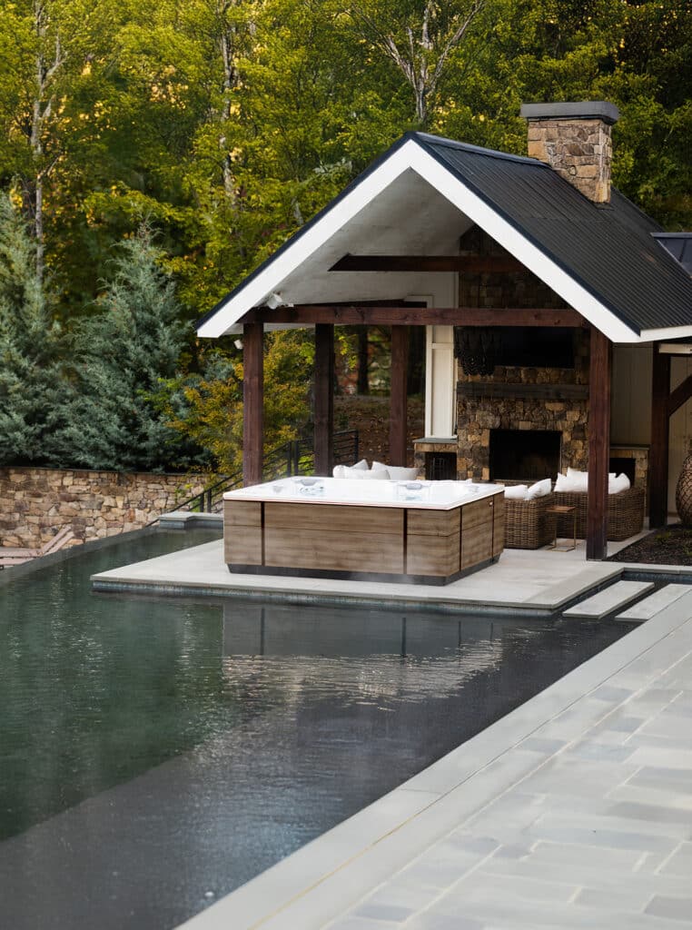 Ruha Stelzmüller zeigt ein Whirlpool, das auf einer Terrasse vor einer kleinen Hütte im Freien steht und außen mit Holz verkleidet ist.