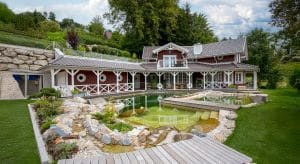 Eine klassische Villa in L-Form mit verspielten Holzelementen, überdachter Rundumterrasse und einem Schwimmteich mit Steg von SCE Reisinger Bau.