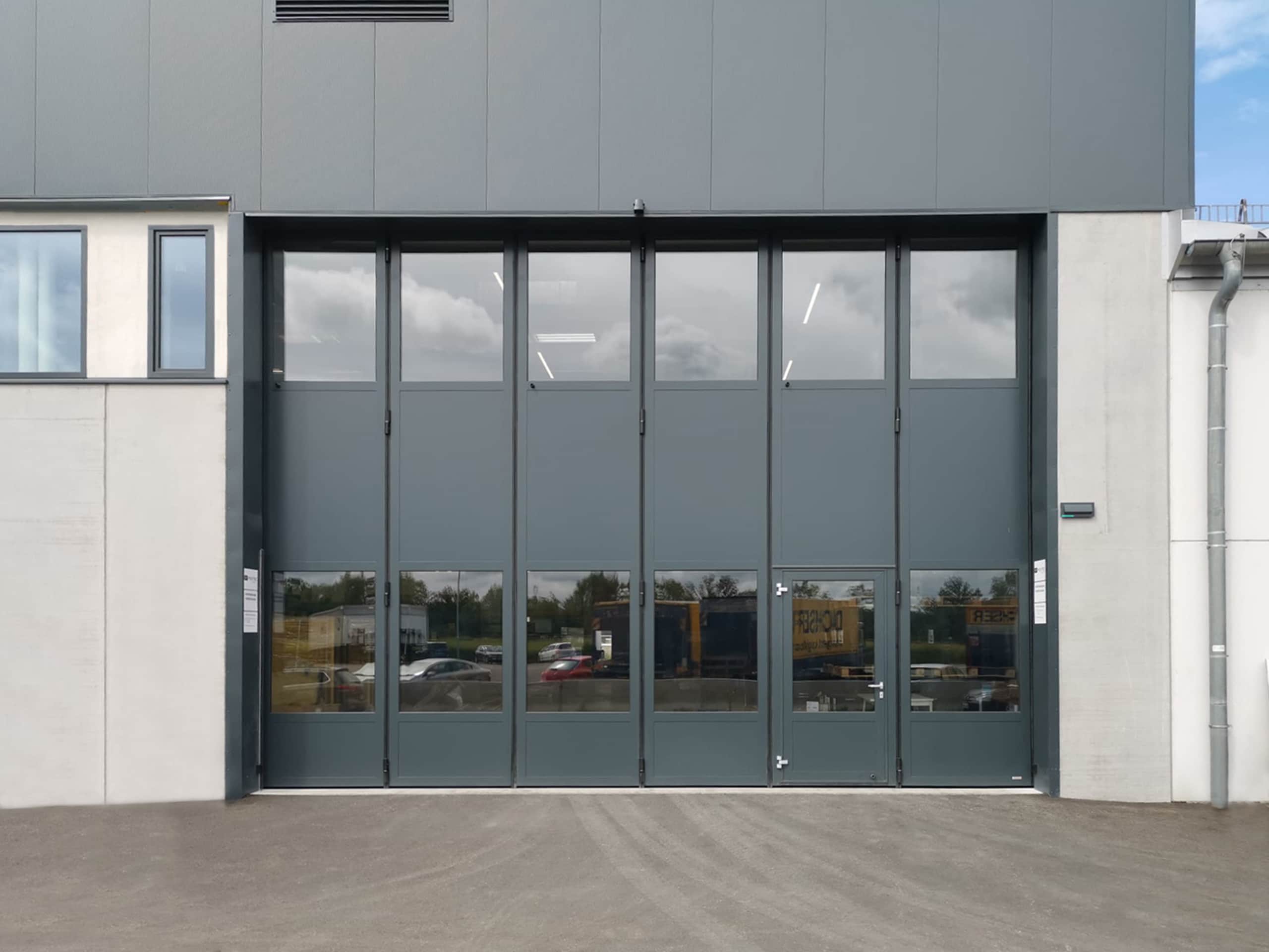 SCHNEIDER Torsysteme zeigt eine Industriehalle mit einem gewerblichen teils verglasten Falttore in anthrazit von außen, das geschlossen ist.