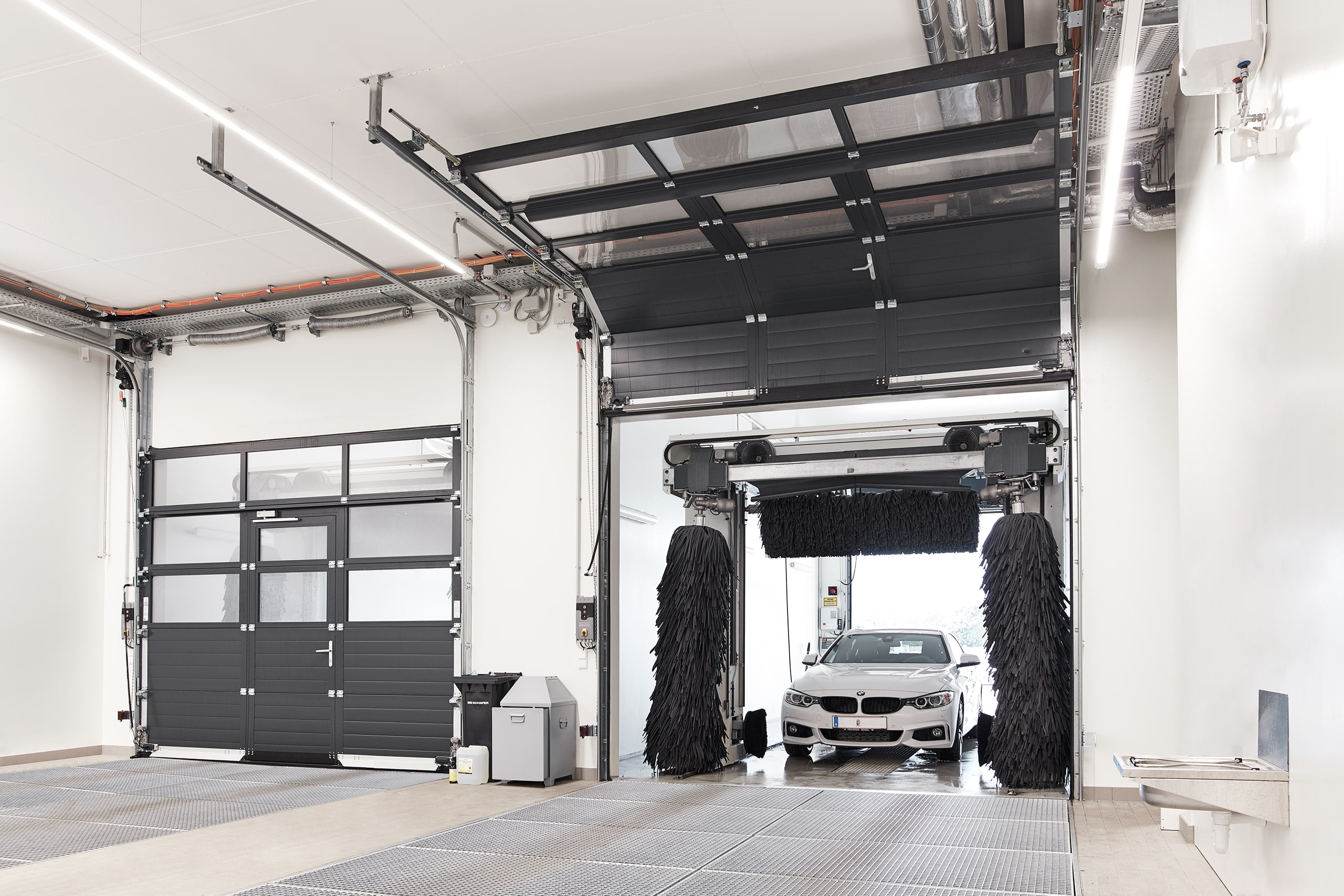 SCHNEIDER Torsysteme zeigt ein gewerbliches Deckensektionaltor einer Waschanlage mit Verglasung im oberen Teil und geschlossenen Elementen im unteren Teil mit integrierter Türe, das gerade geöffnet wird.