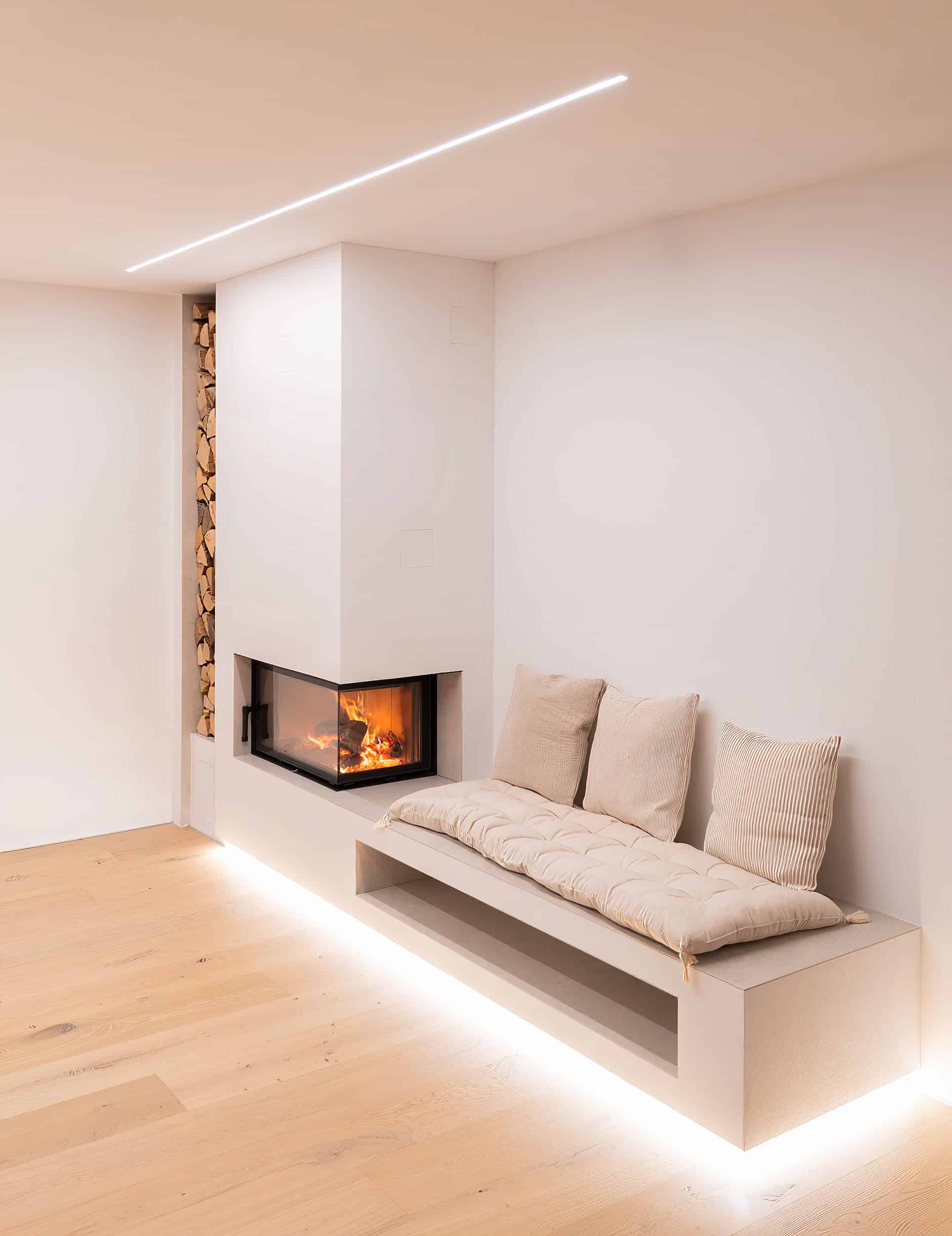 Schlicht eleganter, weiß verputzter Kamin mit Ecksichtfenster, angebauter Sitzbank mit indirekter Bodenbeleuchtung und seitlich integriertem Holzlager von Uwe Seidl Ofenbau.