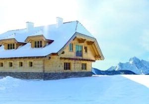 Rustikales schneebedecktes Holzhaus in den Bergen mit klassischen Holzsprossenfenstern, Walmdach, Walmgauben, Schornsteinen und einem Fassadenelement bzw. Fundament aus Stein von SILBER Fensterbau.
